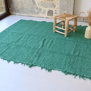 May include: A teal green woven rug with frayed edges and white fringe. A wooden stool with a woven seat sits on the rug.