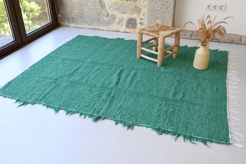 May include: A teal green woven rug with frayed edges and white fringe. A wooden stool with a woven seat sits on the rug.