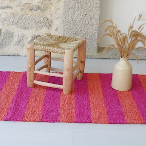 May include: A pink and orange striped rug with a wooden stool with a woven straw seat in the center. A vase with dried grass is on the rug.