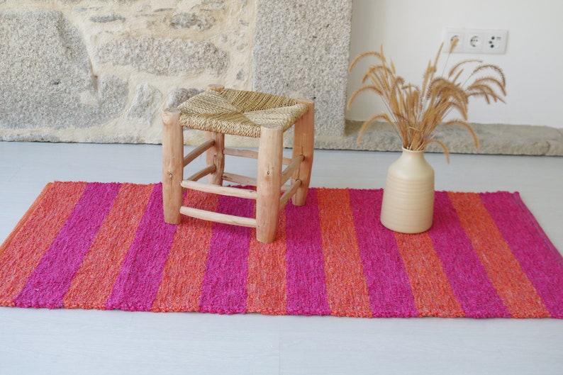 May include: A pink and orange striped rug with a wooden stool with a woven straw seat in the center. A vase with dried grass is on the rug.