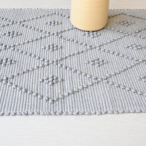 Puede incluir: Una alfombra tejida gris con un patr&oacute;n de diamantes. La alfombra tiene una superficie texturizada con peque&ntilde;os bultos en relieve.