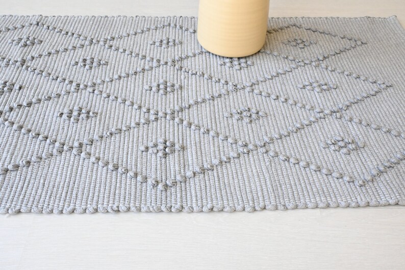 Puede incluir: Una alfombra tejida gris con un patr&oacute;n de diamantes. La alfombra tiene una superficie texturizada con peque&ntilde;os bultos en relieve.