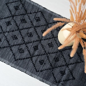 Può includere: Un tappeto tessuto grigio scuro con un motivo a rombi. Il tappeto ha una superficie strutturata e un design geometrico. Un vaso bianco con steli di grano secchi è sul lato destro.