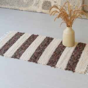 May include: A striped rug with cream and brown bands. The rug has a fringed edge and is on a light grey surface. A beige vase with dried plants sits on the rug. The rug is approximately 100cm long.