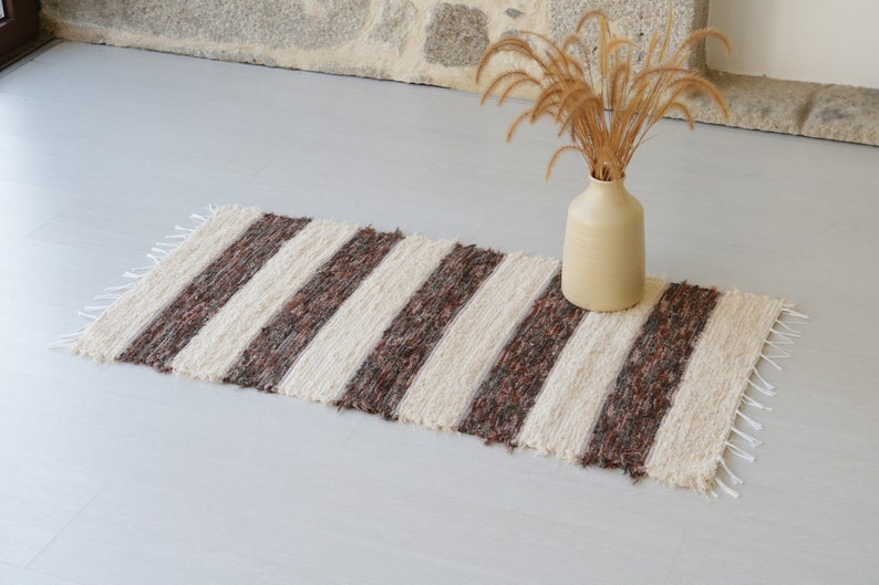 May include: A striped rug with cream and brown bands. The rug has a fringed edge and is on a light grey surface. A beige vase with dried plants sits on the rug. The rug is approximately 100cm long.