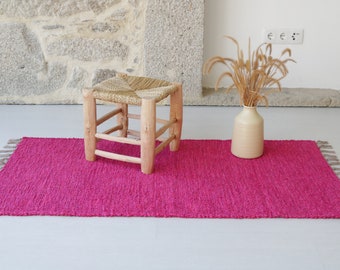 Può includere: Un tappeto rosa tessuto con frange sui bordi e uno sgabello in legno con una seduta intrecciata.