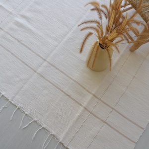 Puede incluir: Una alfombra blanca tejida con flecos en los bordes y un sutil patr&oacute;n a rayas. La alfombra est&aacute; colocada sobre un suelo gris claro con una silla de madera y un jarr&oacute;n de hierba seca en el fondo.