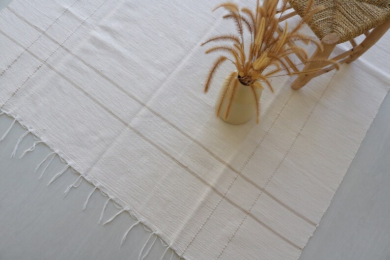 Puede incluir: Una alfombra blanca tejida con flecos en los bordes y un sutil patr&oacute;n a rayas. La alfombra est&aacute; colocada sobre un suelo gris claro con una silla de madera y un jarr&oacute;n de hierba seca en el fondo.