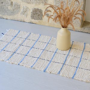Può includere: Un tappeto rettangolare intrecciato con una base color crema, caratterizzato da strisce verticali azzurre e piccoli accenti blu scuro. Il tappeto presenta una superficie testurizzata e corte frange sui due lati corti, creando un semplice motivo a griglia.