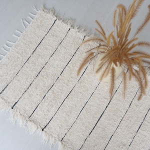 Può includere: Un tappeto bianco e nero a strisce con un bordo sfrangiato. Il tappeto è realizzato in un materiale morbido e soffice e ha un aspetto naturale e organico.