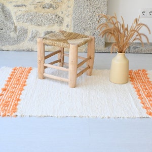 Op de afbeelding: Een wit geweven tapijt met oranje franjes. Een houten kruk met een geweven zitting staat op het tapijt.