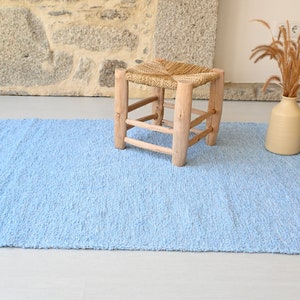 Pu&ograve; includere: Un tappeto shaggy blu chiaro con frange sui bordi. Uno sgabello in legno con una seduta intrecciata si trova sul tappeto.