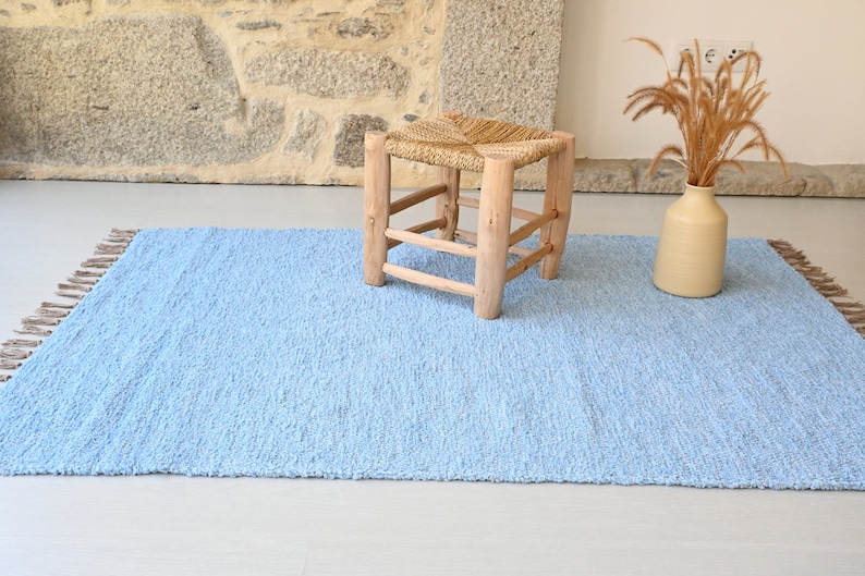 Pu&ograve; includere: Un tappeto shaggy blu chiaro con frange sui bordi. Uno sgabello in legno con una seduta intrecciata si trova sul tappeto.
