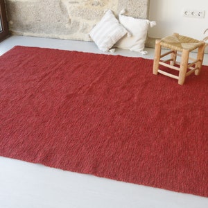 May include: A red, textured rug with a woven design. The rug is laid out on a white floor with a wooden stool and a vase with dried flowers.