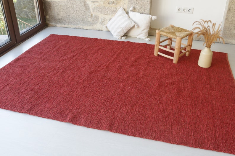 May include: A red, textured rug with a woven design. The rug is laid out on a white floor with a wooden stool and a vase with dried flowers.