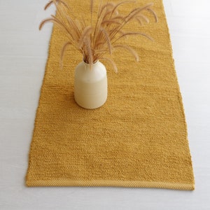 May include: A mustard yellow woven rug with a white ceramic vase filled with dried grass.