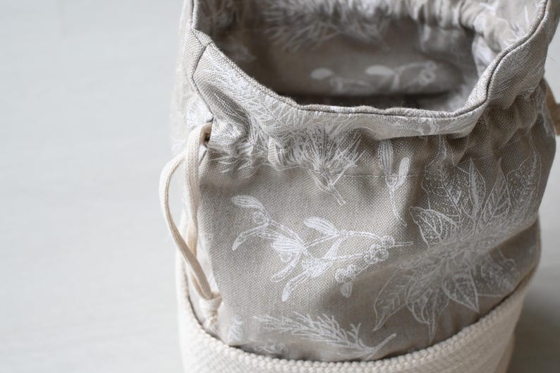 Pu&ograve; includere: Primo piano di un sacchetto di stoffa grigia con coulisse e motivo floreale bianco. L'interno e l'esterno della borsa sono visibili, mostrando il design dettagliato. Il fondo della borsa &egrave; in materiale tessuto bianco sporco, con un cordoncino.