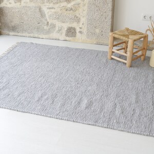 May include: A gray woven rug with fringe on the edges. The rug is laid out on a white floor with a wooden stool in the background.