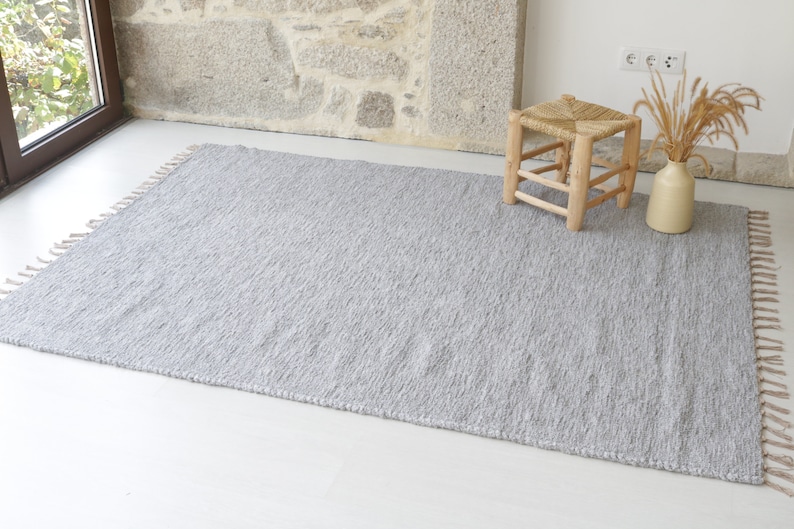 May include: A gray woven rug with fringe on the edges. The rug is laid out on a white floor with a wooden stool in the background.