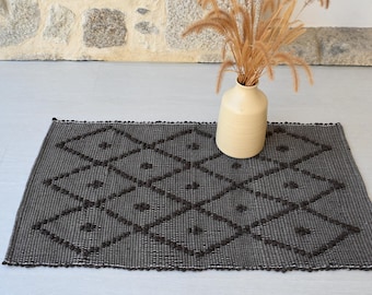 Puede incluir: Una alfombra tejida de color gris oscuro con un patrón de diamantes. La alfombra tiene un borde de flecos y está hecha de un material texturizado.