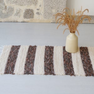 May include: A striped rug in cream and brown, with a fringed edge. The rug is placed on a light grey surface. A beige vase with dried plants is positioned nearby. The rug is suitable for a hallway.