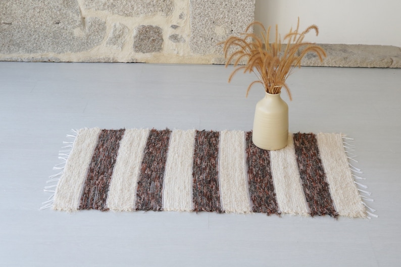 May include: A striped rug in cream and brown, with a fringed edge. The rug is placed on a light grey surface. A beige vase with dried plants is positioned nearby. The rug is suitable for a hallway.