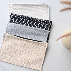 Può includere: Quattro pochette rettangolari intrecciate con cerniera in crema, bianco e nero, grigio e beige. Le pochette sono impilate su una superficie grigio chiaro. Un vaso con piante secche è sullo sfondo.