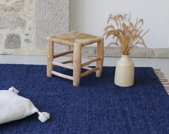 Puede incluir: Una alfombra azul con textura tejida y bordes con flecos. Un pequeño taburete de madera con un asiento tejido se encuentra sobre la alfombra. Una almohada blanca con borlas se encuentra sobre la alfombra. Un jarrón con hierba seca se encuentra sobre la alfombra.
