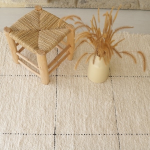 May include: A white woven rug with black dotted lines, a small wooden stool with a woven seat, and a vase with dried grass.