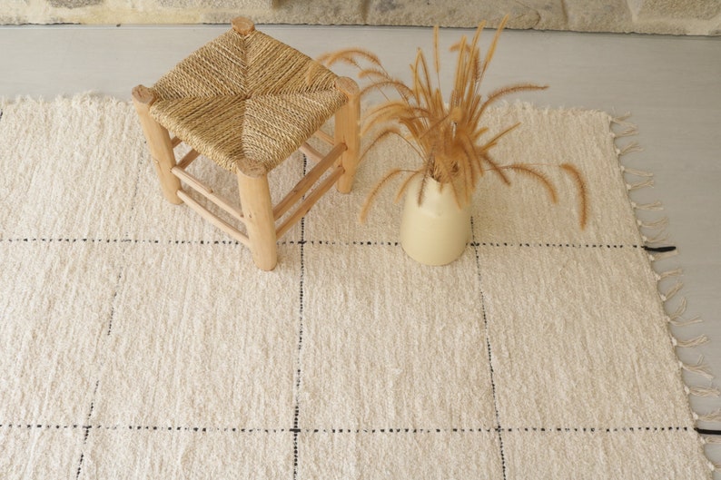 May include: A white woven rug with black dotted lines, a small wooden stool with a woven seat, and a vase with dried grass.