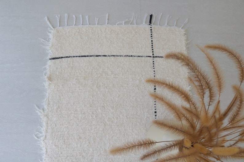 May include: A white rug with black lines forming a grid pattern. The rug has a fringe border and is lying on a light gray surface. A bouquet of dried brown grass is in the lower right corner of the image.