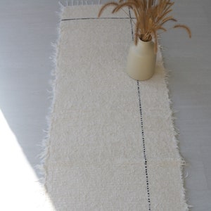 May include: A white rug with a black stitched line down the center. A vase with dried grass is on the rug.