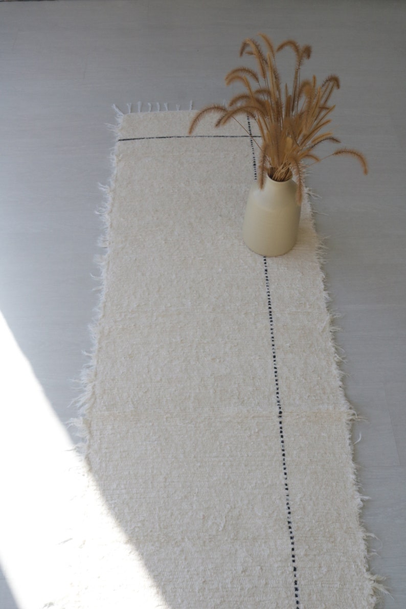 May include: A white rug with a black stitched line down the center. A vase with dried grass is on the rug.
