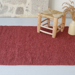 May include: A red woven rug with fringed edges. A wooden stool with a woven seat sits on the rug.
