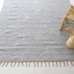 May include: A gray woven rug with fringe on the edges. The rug is on a white surface and a small wooden stool with a woven seat is in the upper right corner of the image.