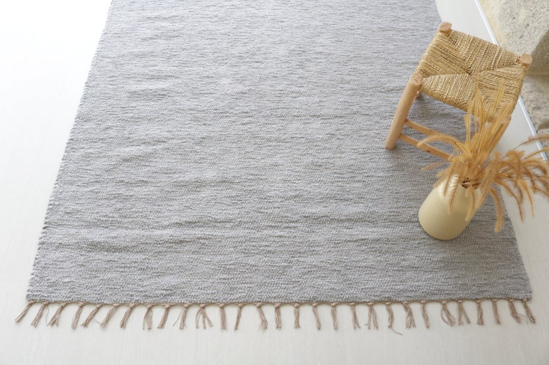 May include: A gray woven rug with fringe on the edges. The rug is on a white surface and a small wooden stool with a woven seat is in the upper right corner of the image.