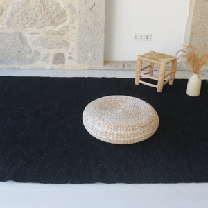 May include: A black woven rug with a round, white woven cushion in the center. A small wooden stool with a woven seat is in the background.
