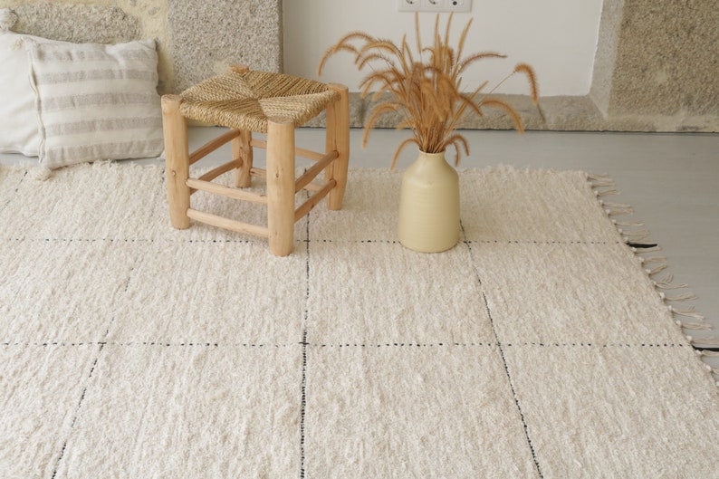 May include: A white woven rug with a subtle grid pattern and fringed edges. A wooden stool with a woven seat sits on the rug, and a vase with dried grasses is in the background.