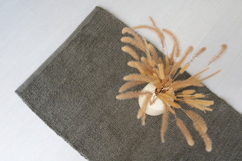 May include: A dark gray rug with a textured surface. A vase with dried beige grass is placed on the rug.