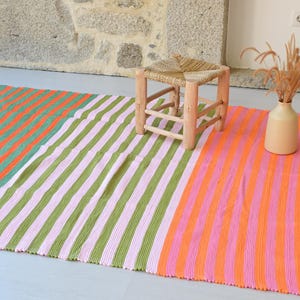 Op de afbeelding: Een rechthoekige geweven plaid met verticale strepen in afwisselende kleuren oranje, groen en roze. De deken heeft witte franjes aan de korte zijden en wordt getoond met een kleine houten kruk en een beige vaas met gedroogd gras.