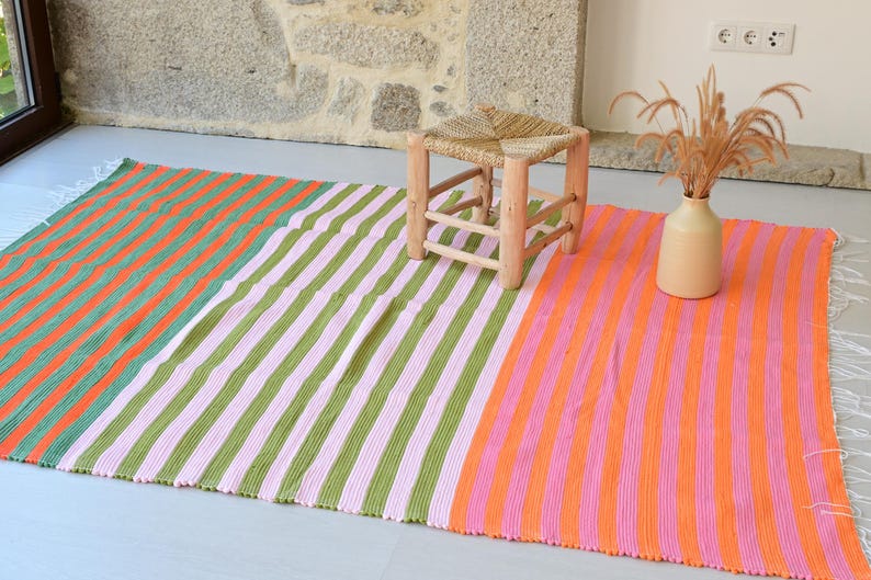 Op de afbeelding: Een rechthoekige geweven plaid met verticale strepen in afwisselende kleuren oranje, groen en roze. De deken heeft witte franjes aan de korte zijden en wordt getoond met een kleine houten kruk en een beige vaas met gedroogd gras.