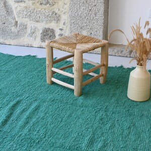 May include: A green woven rug with a natural woven stool and a vase with dried grass.