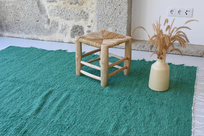 May include: A green woven rug with a natural woven stool and a vase with dried grass.