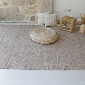 May include: A light brown woven rug with a fringe border. A round woven ottoman and a small wooden stool are on the rug. The rug is in a room with a stone wall and white walls.