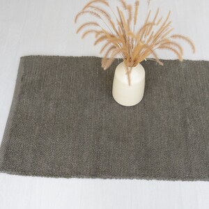 May include: A dark gray woven rug with a light brown vase and dried flowers on top.