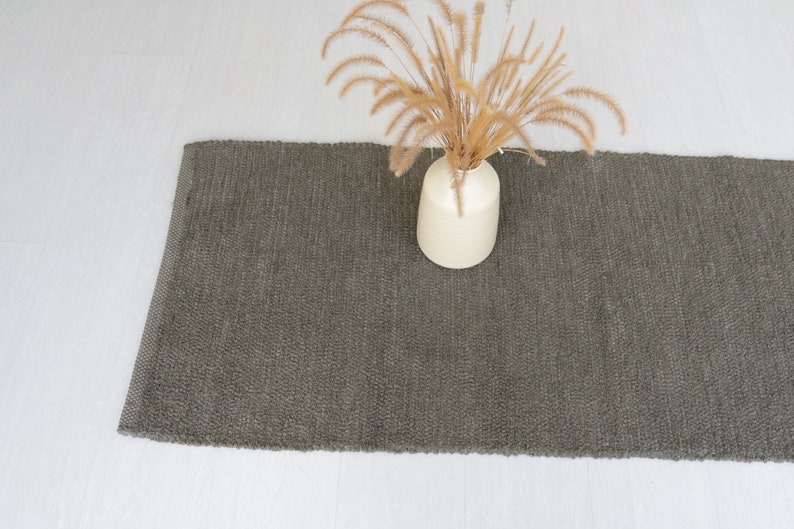 May include: A dark gray woven rug with a light brown vase and dried flowers on top.