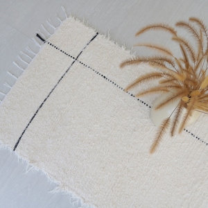 May include: A white rug with black lines and a fringe border. A bouquet of dried brown grass is on top of the rug.