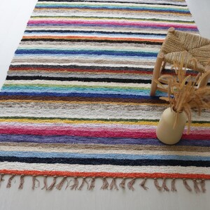 Puede incluir: Una alfombra de rayas coloridas con flecos en los bordes. Un peque&ntilde;o taburete de madera con un asiento tejido y un jarr&oacute;n con flores secas est&aacute;n sobre la alfombra.
