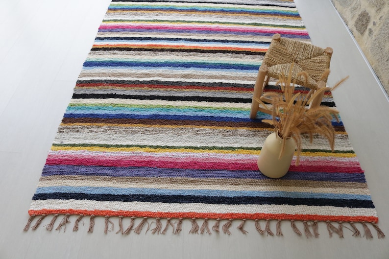 Puede incluir: Una alfombra de rayas coloridas con flecos en los bordes. Un peque&ntilde;o taburete de madera con un asiento tejido y un jarr&oacute;n con flores secas est&aacute;n sobre la alfombra.