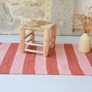 May include: A rectangular rug featuring alternating vertical stripes in shades of terracotta and light pink. A small wooden stool with a woven straw seat sits on the rug, accompanied by a beige ceramic vase containing dried ornamental grasses.
